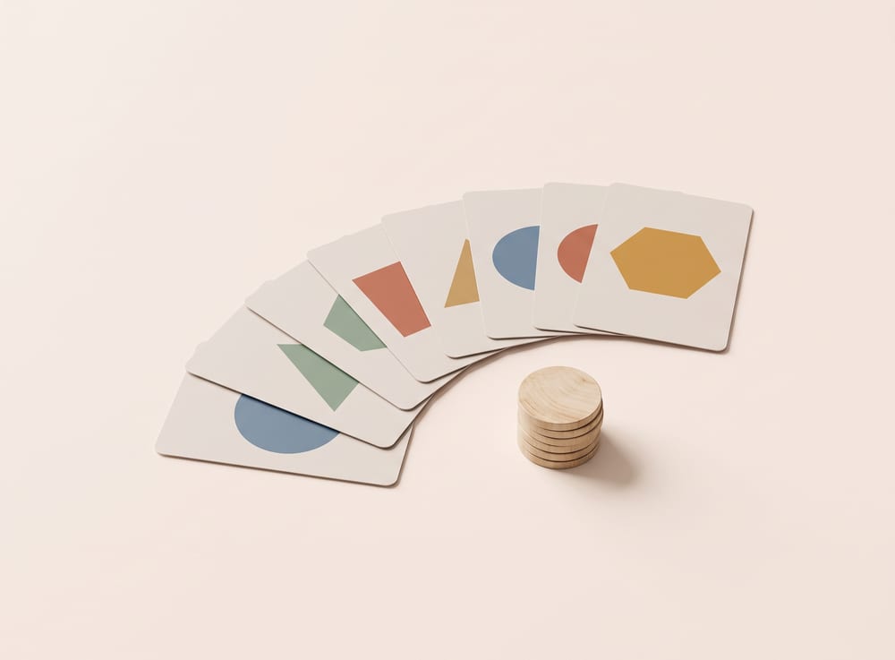 Game cards fanned out in an arc, each carrying a different coloured geometric shape, beside a stack of wooden tokens.