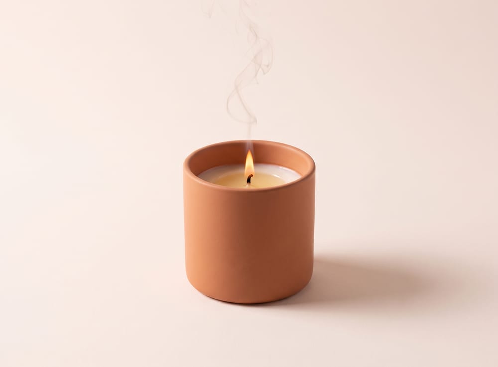 A lit candle in a warm clay ceramic vessel, a thin ribbon of smoke rising.